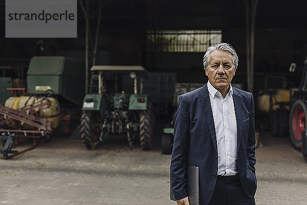 Porträt eines älteren Geschäftsmannes auf einem Bauernhof mit Traktor in einer Scheune