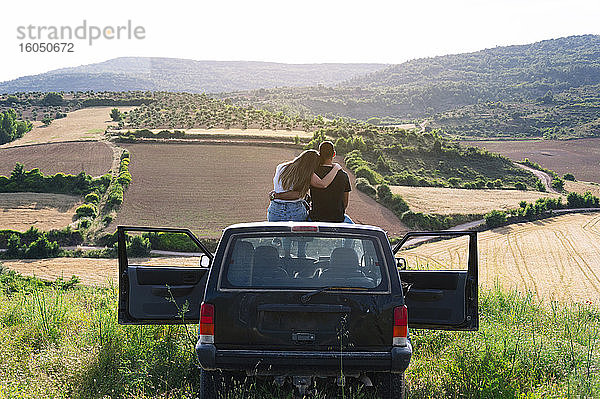 Mid adult couple sitting on 4x4 vehicle roof while looking at landscape during road trip