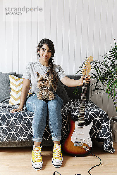 Glückliche junge Frau  die eine E-Gitarre in der Hand hält  während sie mit ihrem Yorkshire Terrier auf dem Sofa zu Hause sitzt