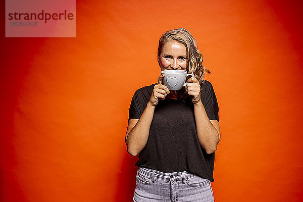 Glückliche Frau hält Kaffeetasse mit Herzform  während sie vor orangefarbenem Hintergrund steht