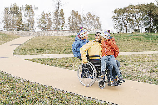 Verspielter Mann mit aufgeregten Söhnen  die im Rollstuhl auf einem Fußweg im Park fahren