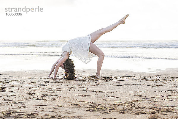 Junge Frau in weißem Kleid tanzt auf Sand am Strand gegen klaren Himmel