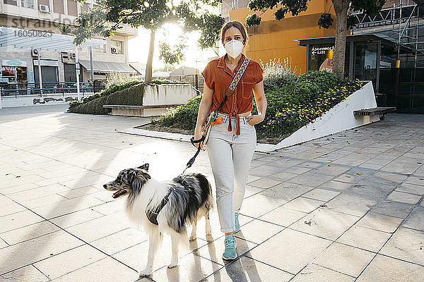 Junge Frau mit Schutzmaske beim Spaziergang mit ihrem Hund in der Stadt