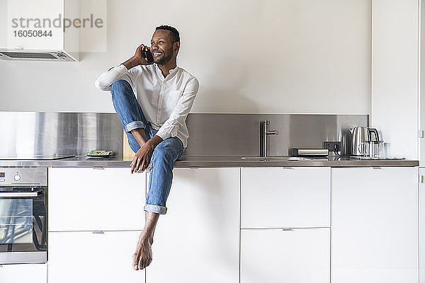 Porträt eines lächelnden Mannes am Telefon  der zu Hause auf dem Küchentisch sitzt und in die Ferne schaut
