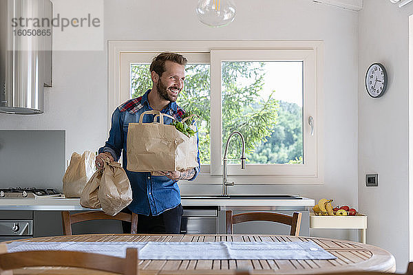 Lächelnder gutaussehender Mann  der eine Tüte mit Lebensmitteln hält  während er in der Küche zu Hause wegschaut