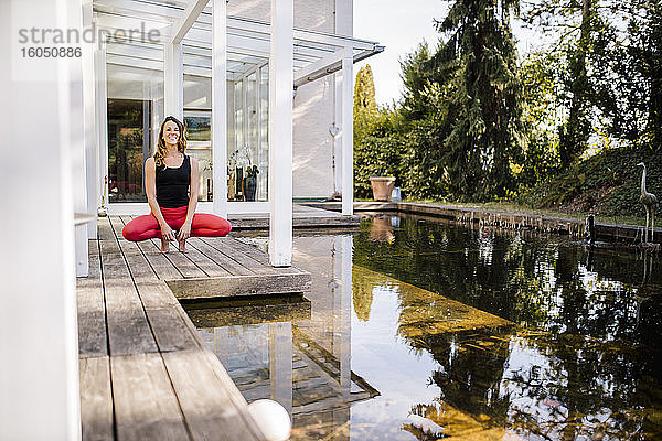 Mittlere erwachsene Frau meditiert hockend auf Hartholzboden am Teich