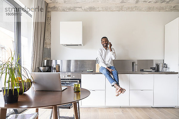 Porträt eines lächelnden Mannes am Telefon  der auf dem Küchentisch in einer modernen Wohnung sitzt