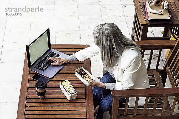 Geschäftsfrau mit grauem Haar  die einen Laptop benutzt  während sie im Garten zu Mittag isst