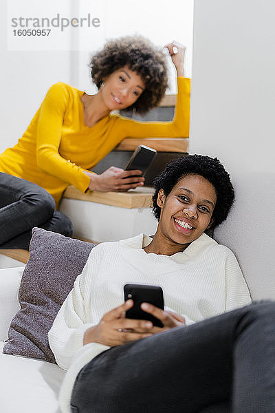 Porträt einer glücklichen jungen Frau  die sich mit ihrem Smartphone auf der Couch entspannt  während ihr Freund im Hintergrund steht