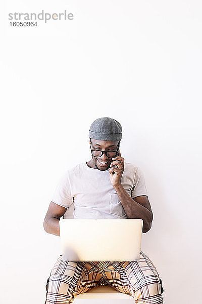 Lächelnder junger Mann  der mit dem Handy telefoniert und einen Laptop vor weißem Hintergrund benutzt