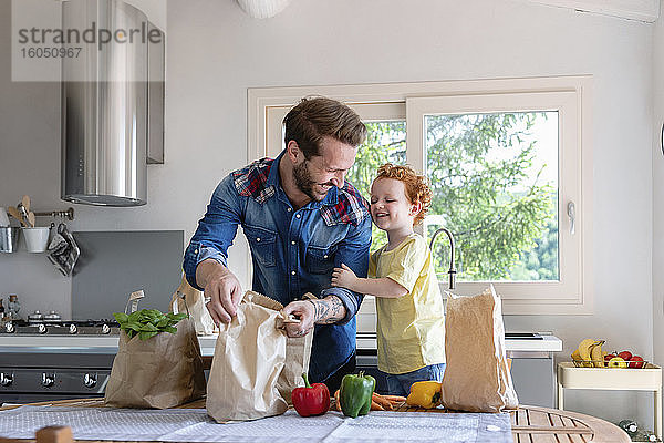Fröhlicher Vater und Sohn mit Lebensmitteln am Esstisch in der Küche stehend