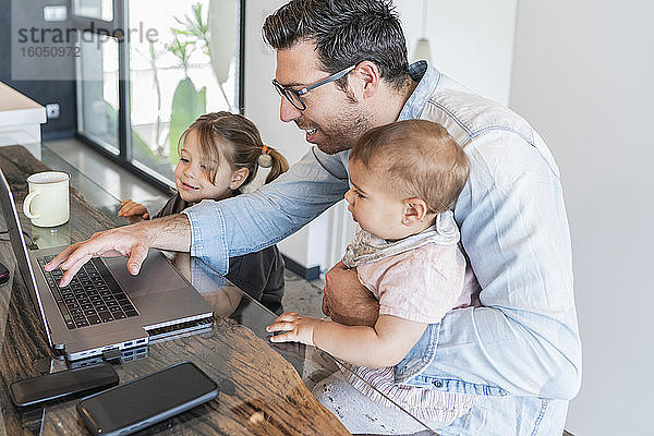 Vater arbeitet über Laptop auf Tisch mit Töchtern an seiner Seite zu Hause