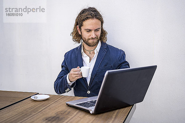 Junger männlicher Unternehmer  der Kaffee trinkt und einen Laptop an der weißen Wand eines Cafés benutzt