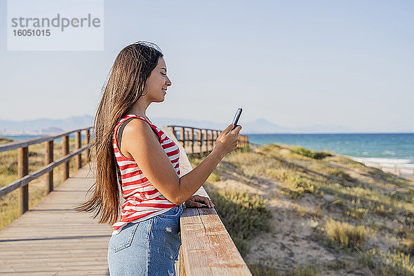 Teenager-Mädchen  das ein Smartphone benutzt  während es an der Strandpromenade vor einem klaren blauen Himmel steht