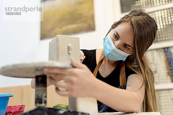 Nahaufnahme einer jungen Frau mit Maske beim Töpfern im Atelier