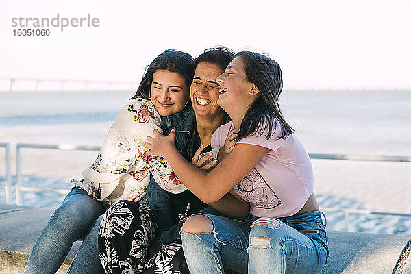 Liebevolle Töchter  die ihre fröhliche Mutter umarmen  während sie auf einer Stützmauer am Meer sitzen