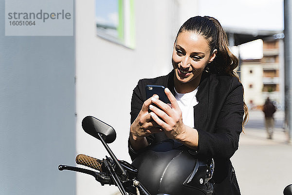 Lächelnde Frau  die auf einem Motorrad sitzt und ein Smartphone benutzt