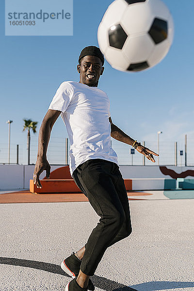 Lächelnder junger Mann spielt Fußball gegen den klaren Himmel im Hof