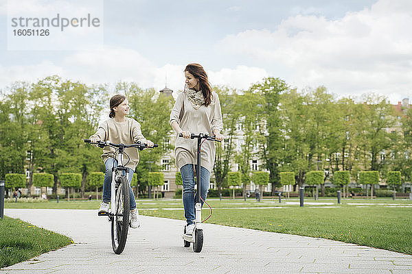 Mädchen fährt Rad  während die Mutter im Stadtpark einen Elektroroller fährt