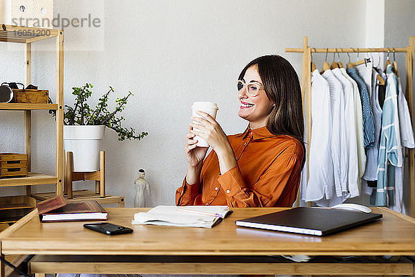 Weibliche Modedesignerin sitzt zu Hause am Schreibtisch und macht eine Kaffeepause