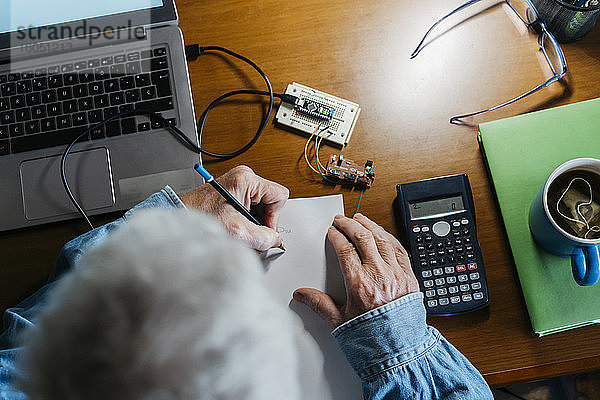 Ausgeschnittenes Bild eines älteren Mannes  der bei der Reparatur eines Laptops zu Hause ein Diagramm auf Papier erstellt