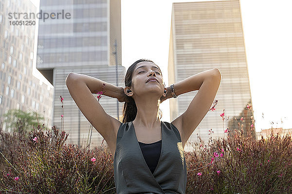 Entspannte junge Geschäftsfrau  die in der Pause mit den Händen hinter dem Kopf vor einem Bürogebäude Musik über Kopfhörer hört