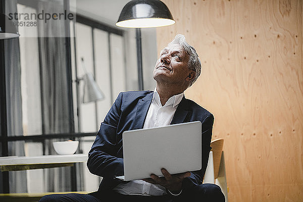 Älterer Geschäftsmann sitzt unter einer Deckenlampe  benutzt einen Laptop und schaut ins Licht