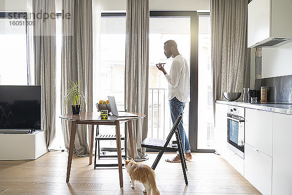 Mann  der in einer modernen Wohnung vor der Balkontür steht und ein Smartphone benutzt