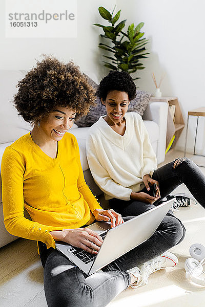 Lächelnde junge Frau  die zu Hause auf dem Boden sitzt und einen Laptop benutzt  während ihre Freundin sie beobachtet