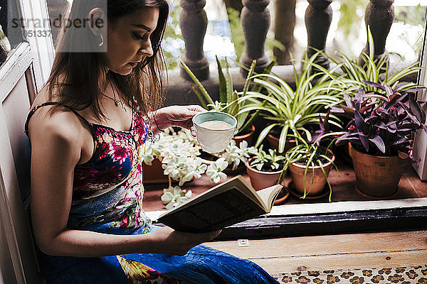 Frau hält Tasse beim Lesen eines Buches auf dem Balkon zu Hause