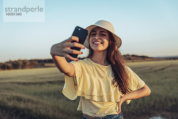 Glückliche junge Frau nimmt Selfie durch Smartphone gegen klaren Himmel bei Sonnenuntergang