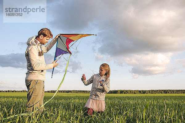 Vater und Tochter lassen einen Drachen steigen  während sie auf einer grünen Landschaft vor dem Himmel stehen