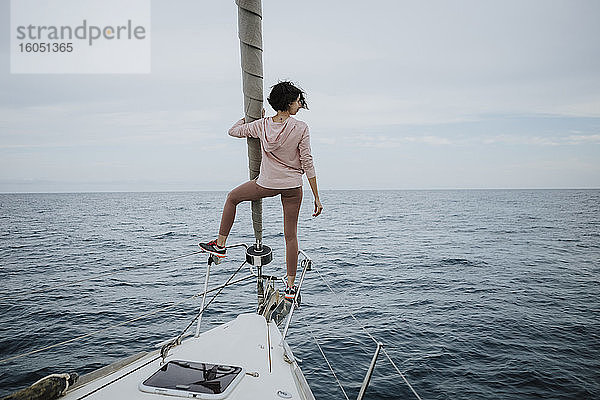 Weiblicher Segler  der auf dem Bug eines Segelboots im Meer steht und die Aussicht betrachtet
