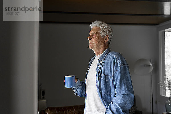 Nachdenklicher älterer Mann  der eine Kaffeetasse hält  während er durch ein Fenster zu Hause schaut