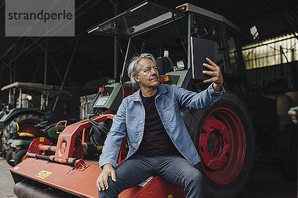 Älterer Mann mit Tablet auf einem Bauernhof mit Traktor in der Scheune