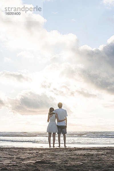 Verliebtes junges Paar  das sich in den Armen liegt und auf das Meer vor einem bewölkten Himmel blickt