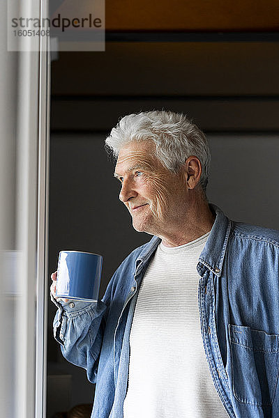 Nachdenklicher älterer Mann lächelt beim Kaffeetrinken am Fenster zu Hause