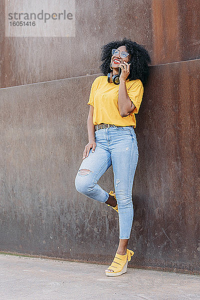 Lächelnde Frau mit Afro-Haar und Brille  die ein Smartphone benutzt und sich an eine Wand lehnt
