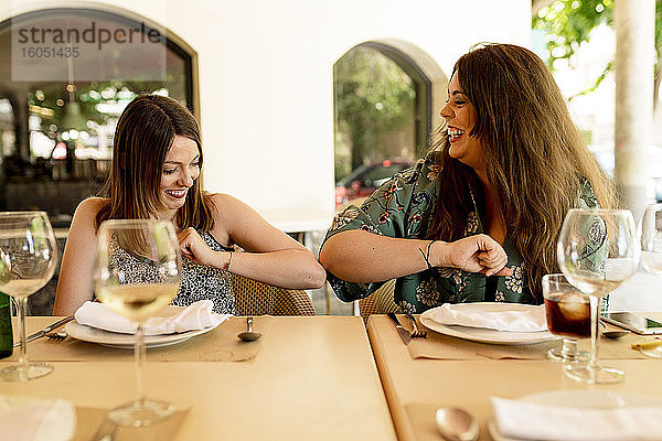 Glückliche junge Frauen grüßen sich mit Ellbogenstößen  während sie am Restauranttisch sitzen