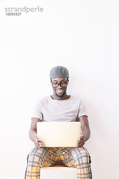 Lächelnder junger Mann sitzt mit Laptop vor weißem Hintergrund