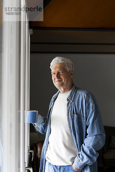 Selbstbewusster älterer Mann  der eine Kaffeetasse hält  während er zu Hause am Fenster steht