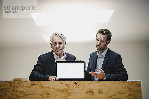 Zwei selbstbewusste Geschäftsleute sitzen in einem Büro  präsentieren einen Laptop-Bildschirm und lächeln.