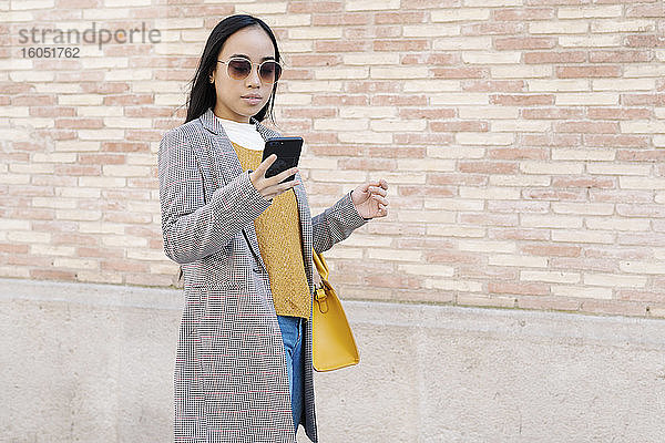Stilvolle junge Frau mit Sonnenbrille  die ein Smartphone benutzt  während sie in der Stadt an der Wand steht