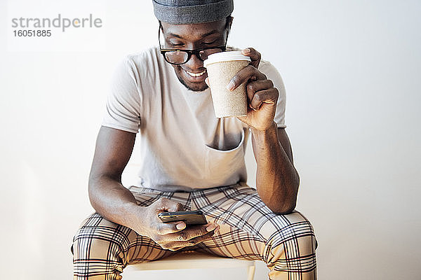 Lächelnder Mann  der eine Textnachricht schreibt  während er eine Kaffeetasse hält und vor einem weißen Hintergrund sitzt