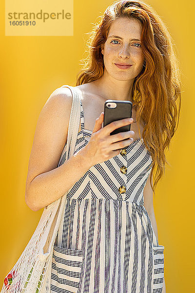 Lächelnde schöne Frau hält Smartphone im Stehen vor gelbem Hintergrund