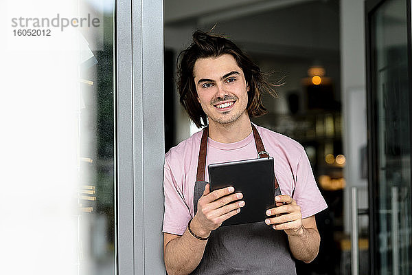 Lächelnder männlicher Besitzer  der ein digitales Tablet benutzt  während er am Eingang eines Cafés steht