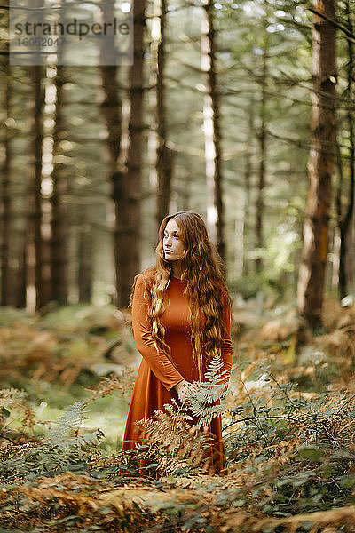 Nachdenkliche schöne Frau mit langem Haar inmitten von Pflanzen im Wald stehend
