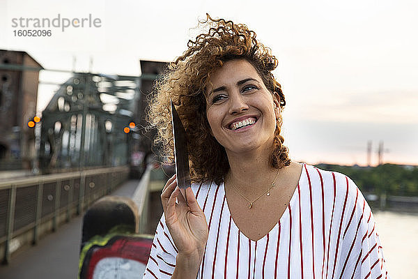 Fröhliche Frau mit lockigem Haar  die über ein digitales Tablet spricht  gegen den Himmel in der Stadt