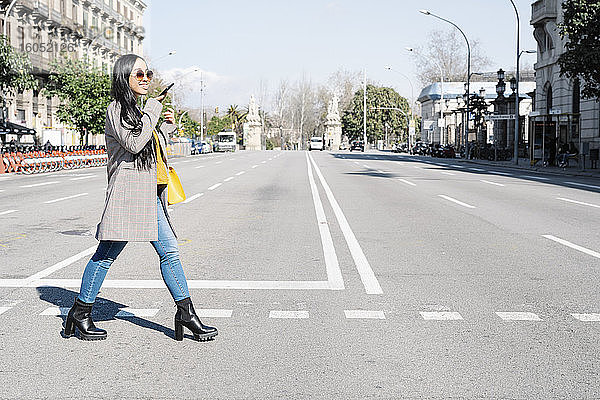 Stilvolle junge Frau spricht über Smartphone beim Überqueren der Straße in der Stadt