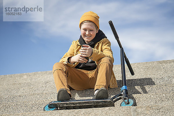 Glücklicher Junge  der sein Smartphone benutzt  während er mit seinem Roller auf einer Treppe gegen den Himmel sitzt